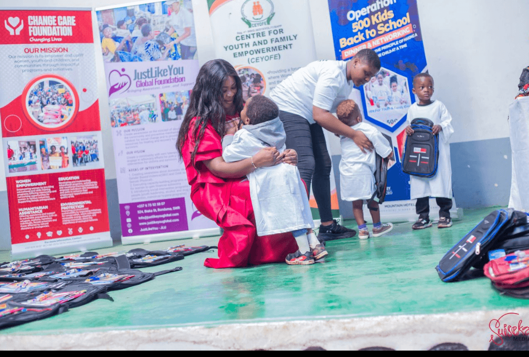 TMTF founder Dorothy Akoh-Arrey kneeling to help a young child try on a school top, with rows of packed backpacks laid out beside them